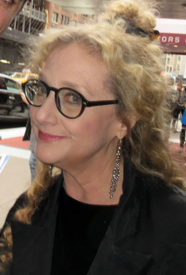 Portrait of a smiling woman with curly blonde hair pinned up, round dark-framed glasses and a long sparkling earring, wearing black, on a city street.