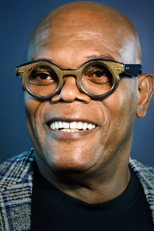 Close portrait of a smiling bald man wearing round gold lattice-patterned glasses and a checked jacket over a black top, against a blue background.