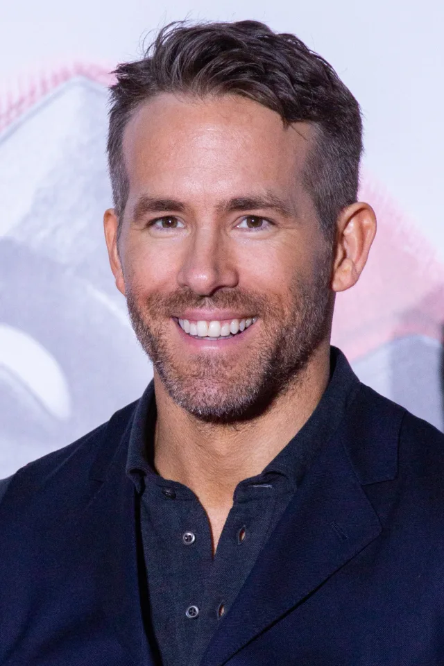 Smiling man with short dark hair and light stubble, wearing a dark navy jacket over a dark buttoned shirt, photographed against a pale backdrop.
