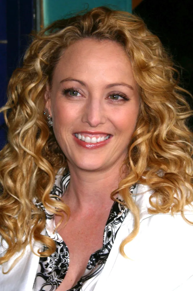 A smiling woman with long, curly blonde hair, wearing a white jacket over a black and white patterned blouse.