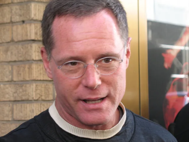 A man with thin round glasses and a dark top with a pale collar, photographed close up outdoors in front of a brick wall.