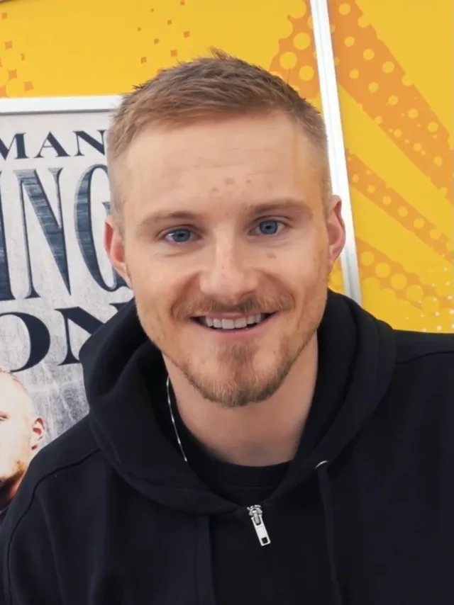 Smiling man with short fair hair and a light beard, wearing a black zip hoodie, seated in front of a bright yellow display.