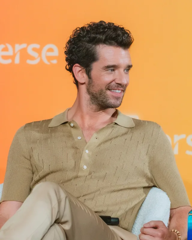 Michael Urie smiling and looking to one side, seated against an orange backdrop, wearing a tan knit polo shirt.