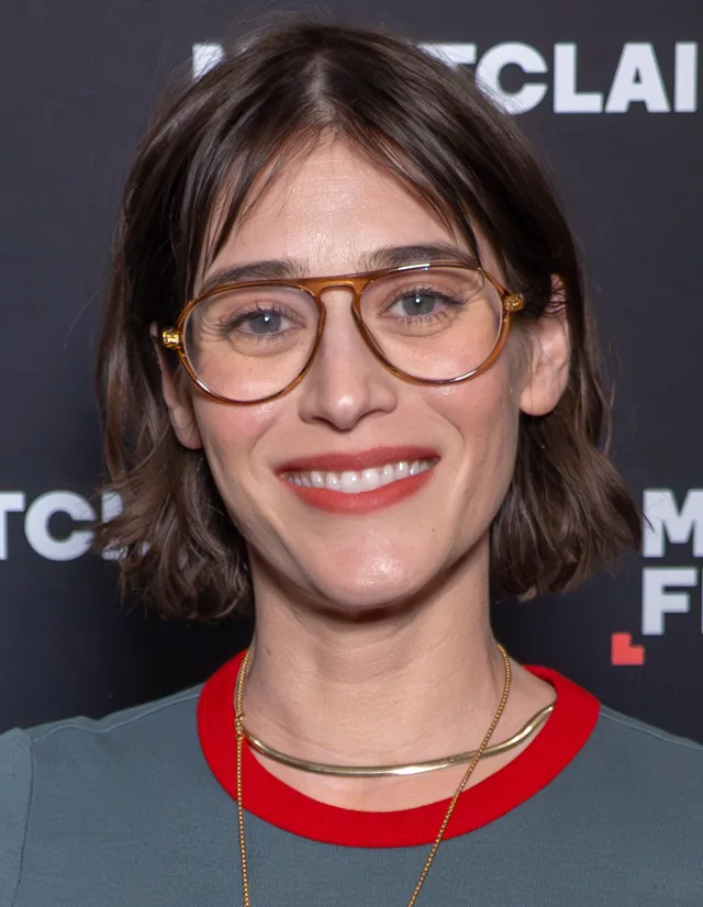 Portrait of a smiling woman with a shoulder-length dark bob and amber aviator glasses, wearing a grey-green top with a red collar and a gold necklace.