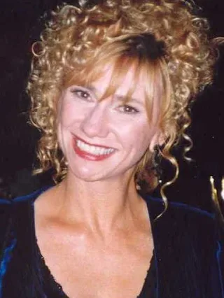 Kathy Baker smiling, with curly blonde hair piled up and a dark velvet top, photographed at night.
