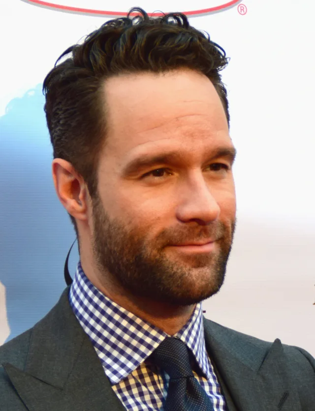 Portrait of Chris Diamantopoulos with dark swept-back hair and a short beard, in a grey jacket and checked shirt with a navy tie.