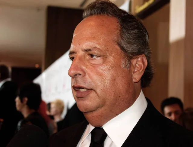 Jon Lovitz in profile at an indoor event, wearing a black jacket, white shirt and black tie, with other guests blurred behind him.