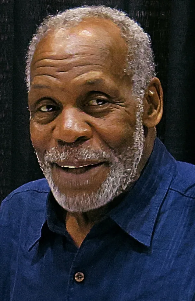 Close photograph of an older man with grey hair and a grey beard, smiling, in a blue collared shirt against a dark curtain.