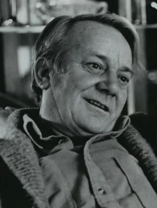 Black and white photograph of an older man with swept-back hair, smiling and looking to one side, wearing a knitted cardigan over an open-collared shirt.