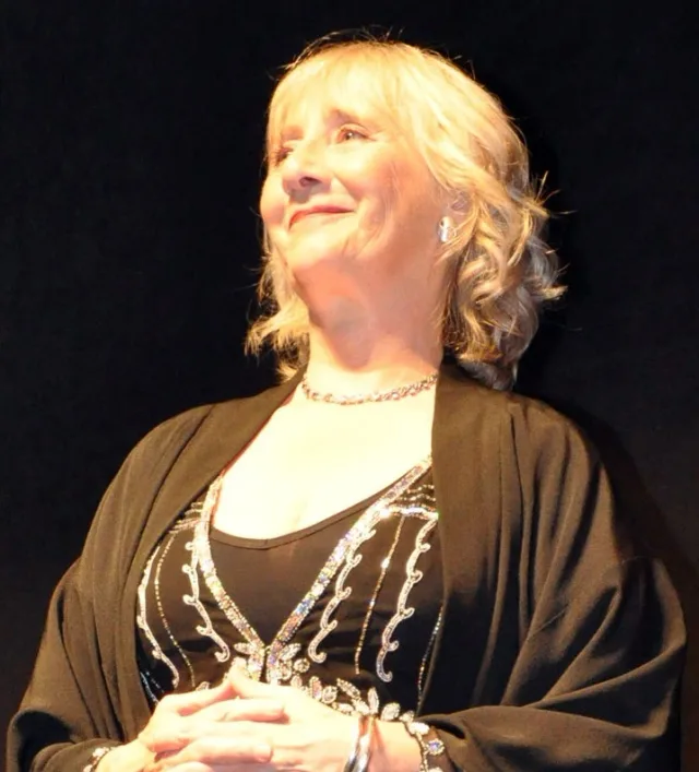 Gemma Jones smiling and looking up to one side, with wavy blonde hair, a sparkling necklace and a dark wrap, against a black background.