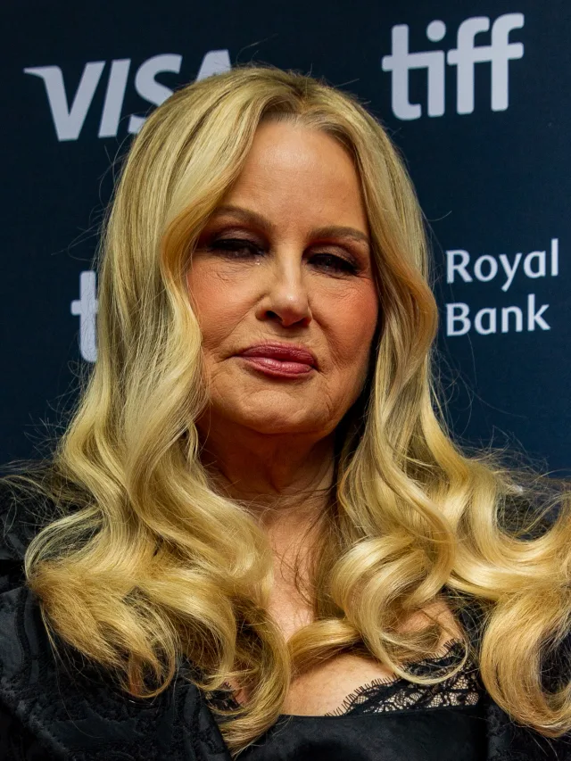 Close portrait of Jennifer Coolidge with long wavy blonde hair, in front of a dark festival backdrop.
