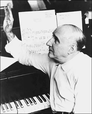 Black and white photograph of a balding older man in a light shirt at a piano, one hand raised, looking at sheet music on the stand.
