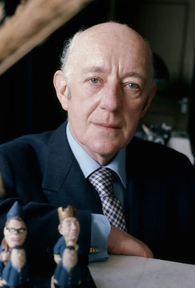 Colour portrait of an older, balding man with pale blue eyes in a navy jacket, blue shirt and checked tie, leaning on a table with two small figurines in front of him.