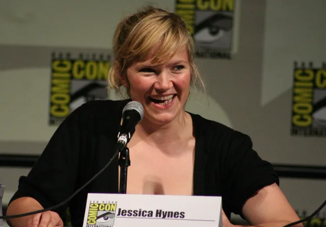 Jessica Hynes laughing at a microphone during a convention panel, her fair hair pinned up, wearing a black cardigan.