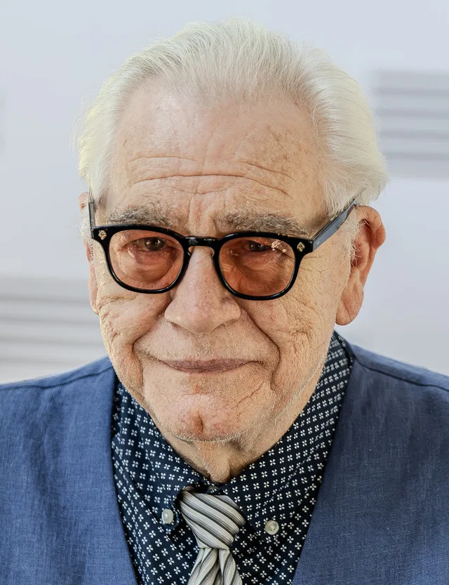 Close portrait of an older man with swept-back white hair, tinted round glasses and a gentle smile, in a blue jacket over a dark patterned shirt and striped tie.