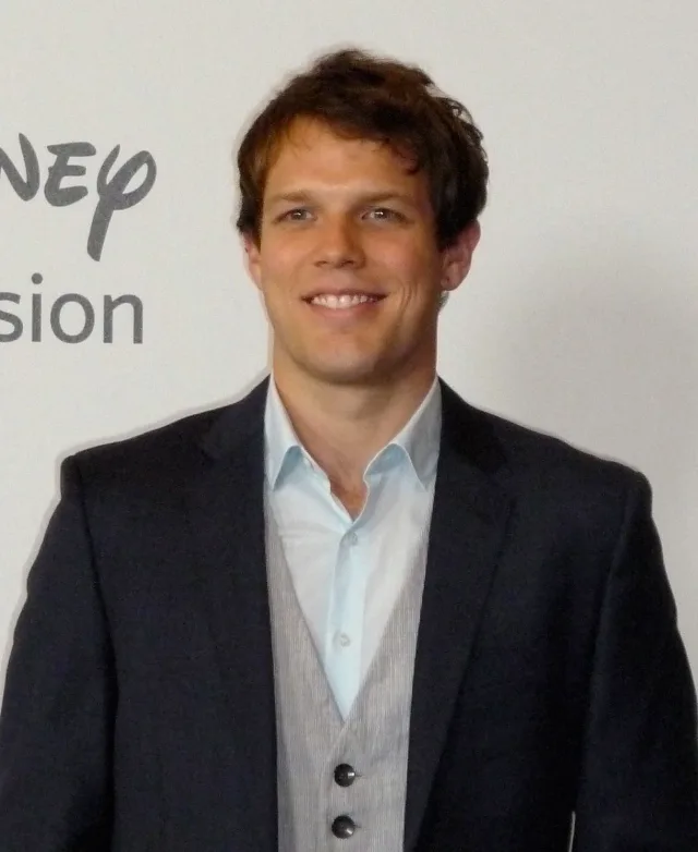 Jake Lacy smiling in a navy blazer, pale blue shirt and grey waistcoat in front of a white backdrop.