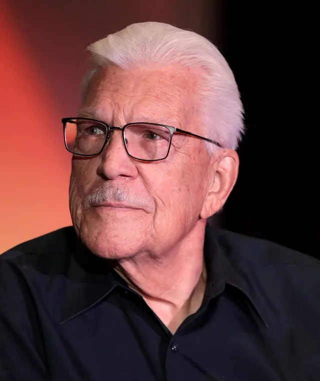 Head and shoulders photograph of an older man with swept-back white hair, glasses and a grey moustache, in a dark shirt.