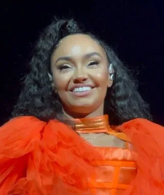 Smiling woman with long dark curly hair in a bright orange ruffled top, photographed against a dark background.
