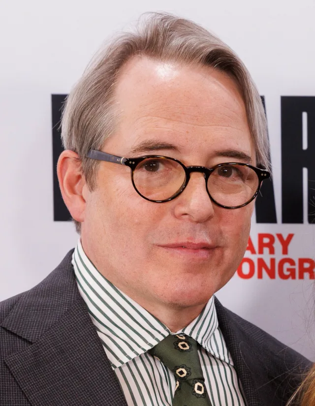 Head and shoulders photograph of Matthew Broderick in round tortoiseshell glasses, a dark jacket and a green striped shirt with a green patterned tie.