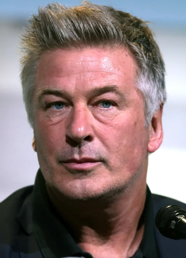 Close portrait of a man with short swept-up grey-blond hair and blue eyes, wearing a dark collared shirt, with the edge of a microphone in the corner.