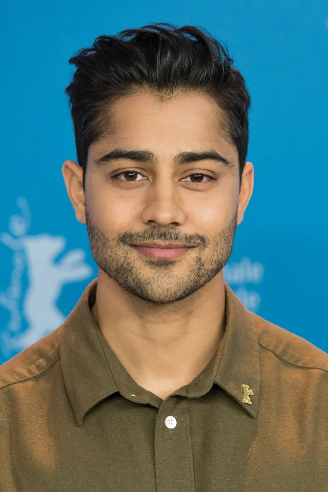 A man with swept-up dark hair and a trimmed beard, wearing an olive collared shirt, looking at the camera against a bright blue backdrop.