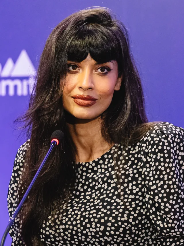 A woman with long dark hair and a fringe, in a black and white spotted top, speaking at a microphone at a press event.