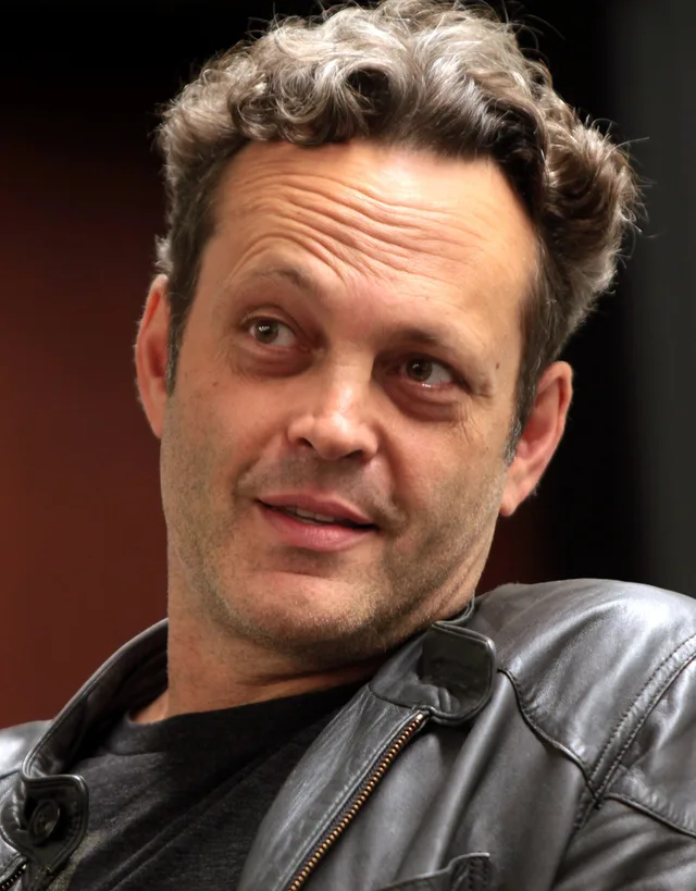 Close-up photograph of a man with curly grey-brown hair and stubble, wearing a dark leather jacket, glancing to one side with a slight smile.