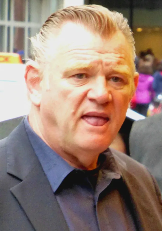 Close photograph of a man with slicked-back fair hair in a dark jacket and blue shirt, speaking outdoors on a city street.