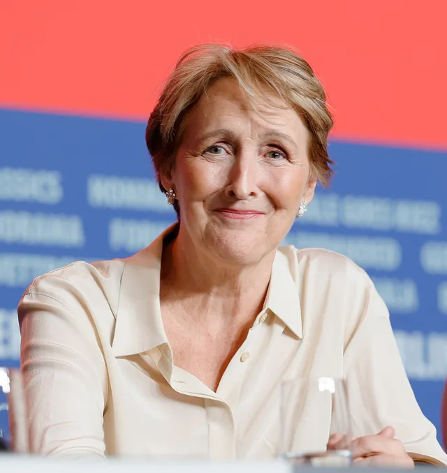 Fiona Shaw at a press conference, smiling slightly, with short fair hair and a cream silk blouse, seated in front of a blue and red backdrop.