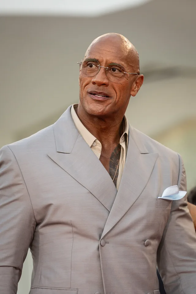 A bald man in thin-rimmed glasses and a light grey double-breasted suit with a pocket square, looking off to one side at an outdoor evening event.