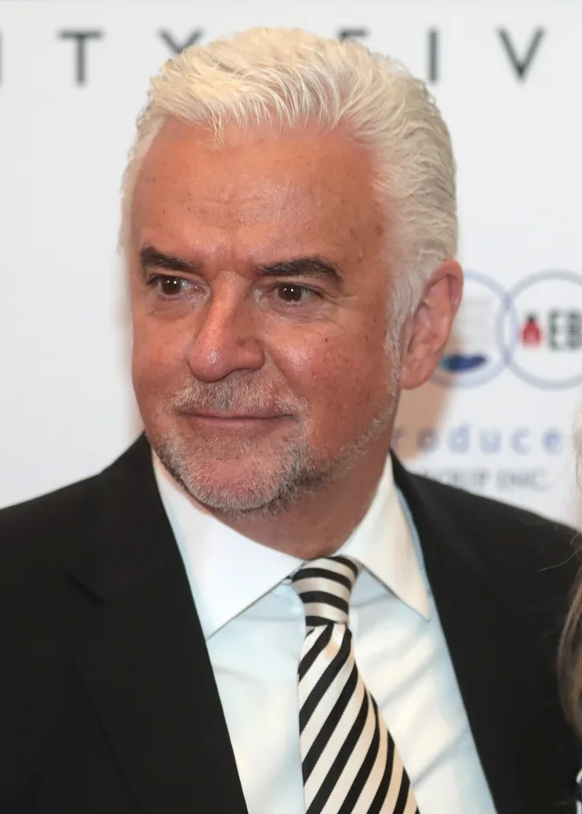 John O'Hurley in close-up, white swept-back hair and a grey beard, in a dark suit and a black and white striped tie.