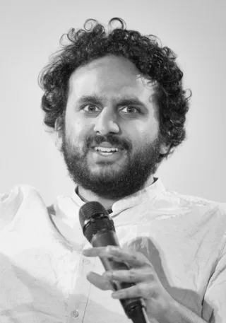 Black and white photograph of a bearded man with dark curly hair speaking into a microphone and gesturing with one hand.