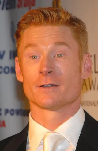 Close portrait of a man with short ginger hair and light eyes, wearing a dark jacket and white shirt and tie, in front of an event backdrop.