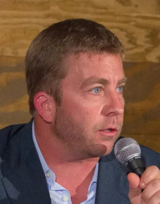 Close photograph of a man with short brown hair and blue eyes, in a navy jacket, speaking into a microphone at a panel.