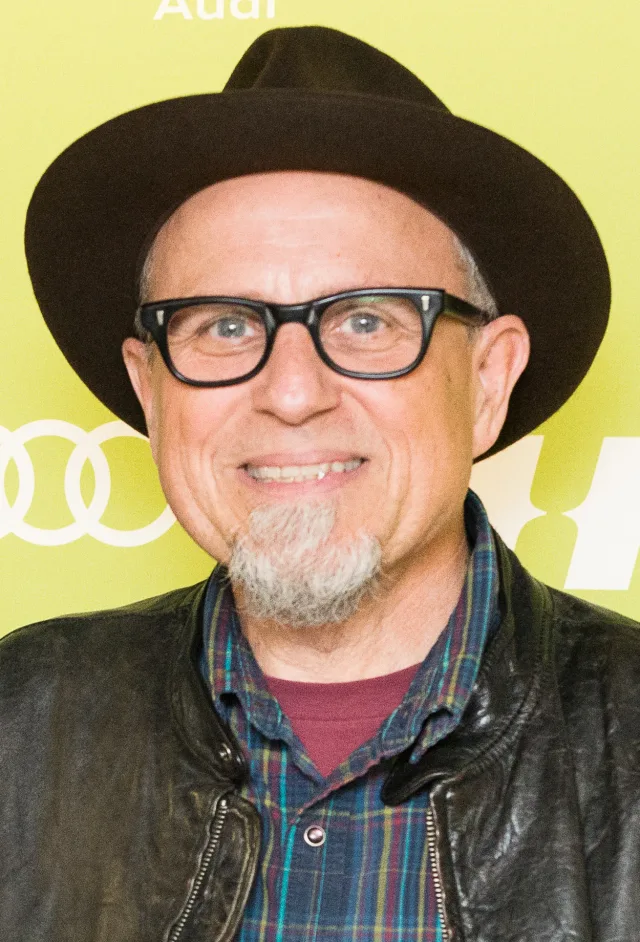 Smiling man in a dark brimmed hat, black-framed glasses and a grey goatee, wearing a leather jacket over a plaid shirt, in front of a yellow-green backdrop.