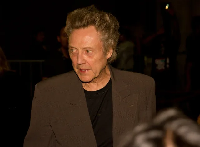 Christopher Walken in a brown jacket and black top, looking to the side in a dim, crowded setting.