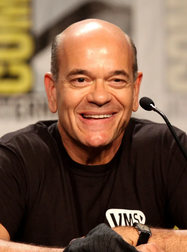 Robert Picardo smiling broadly at a convention panel table, wearing a black t-shirt, with a microphone beside him.