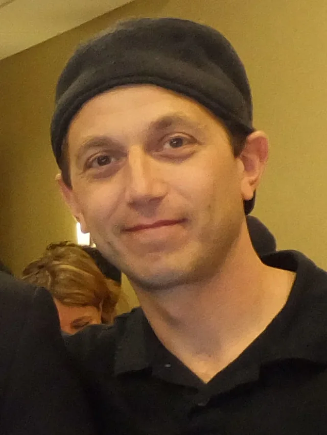 Close photograph of a man with dark eyes wearing a dark flat cap and a black polo shirt, smiling slightly at the camera.