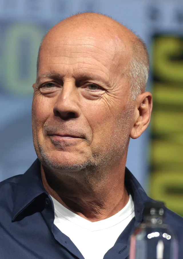 Bruce Willis with a shaved head and light stubble, wearing a navy shirt over a white t-shirt, at a panel event.