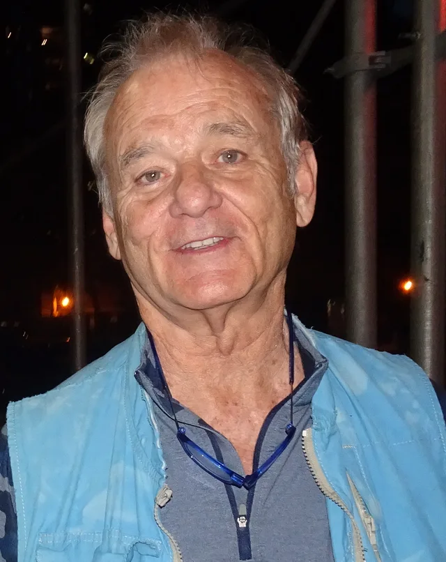 Close photograph of an older Bill Murray smiling, with grey swept hair, wearing a light blue vest over a grey zip-neck top, at night.