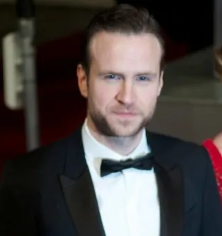 Head and shoulders photograph of a man with short brown hair and light stubble, in a black dinner jacket and bow tie at an evening event.