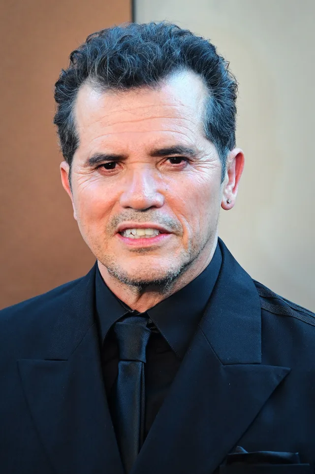 Close portrait of John Leguizamo, a man with short dark curly hair and a grey goatee, wearing a black suit, shirt and tie.