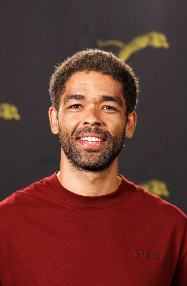 Smiling man with short curly hair and a beard, wearing a maroon sweatshirt, photographed against a dark backdrop.