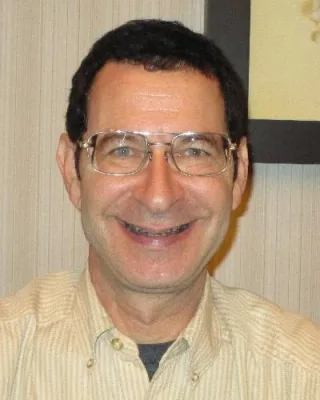 Smiling man with short dark hair and wire-framed glasses, wearing a pale striped collared shirt, indoors against a striped wall.