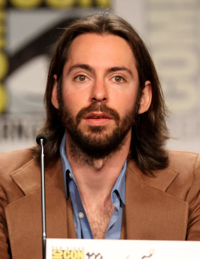 Close portrait of a long-haired, bearded man in a tan jacket and blue shirt speaking at a panel microphone.