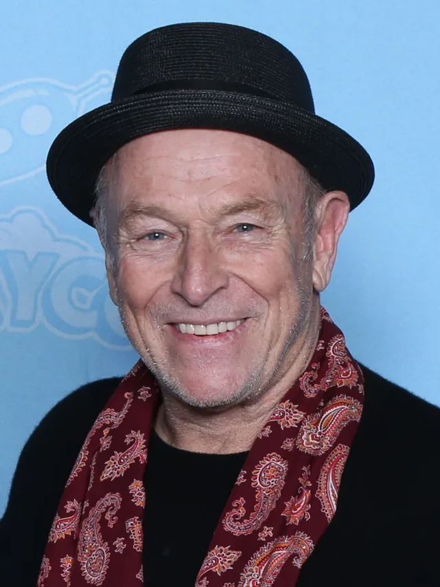 Smiling older man in a black pork-pie hat and a red paisley scarf, photographed against a pale blue backdrop.