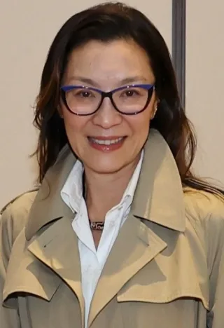 Smiling woman with long dark hair and blue-framed glasses, wearing a beige trench coat over a white shirt.