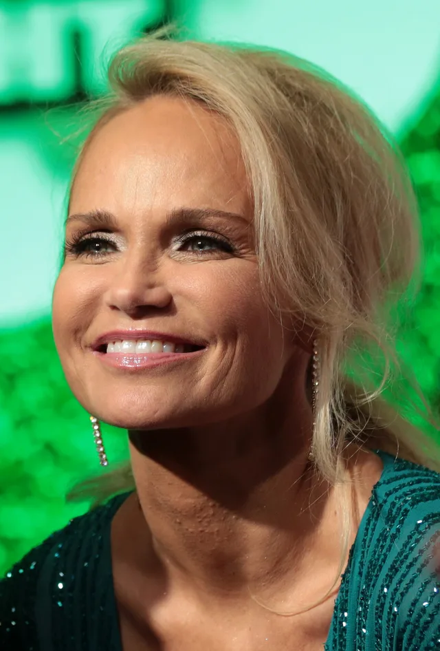 A smiling woman with blonde hair and long drop earrings, wearing a sparkling teal top, in front of a green backdrop.