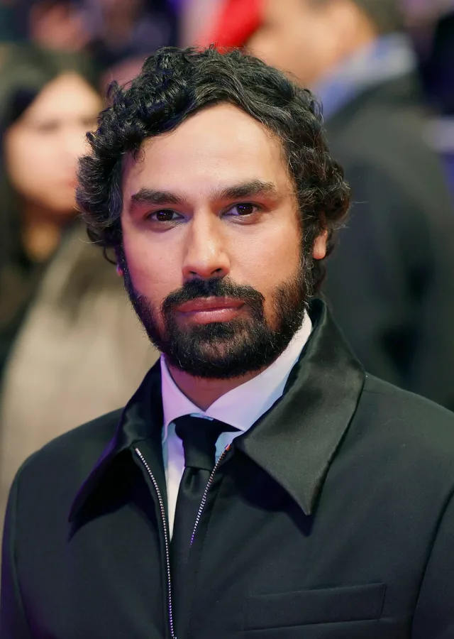 Portrait of a bearded man with dark curly hair, wearing a black zip-collar jacket over a white shirt and black tie, at a red carpet event.