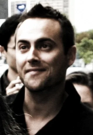 Close portrait of a man with short dark hair and stubble, wearing a dark collared shirt, looking to one side with a slight smile.